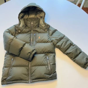 Grön pufferjacka från Polo Ralph Lauren - Snygg mörkgrön pufferjacka från Polo Ralph Lauren med orange logga på bröstet. Jackan har huva, tre dragkedjefickor fram och quiltad design. Perfekt för kalla dagar och har en clean, modern look som funkar till jeans och sneakers.Storlek M kan passa mindre 