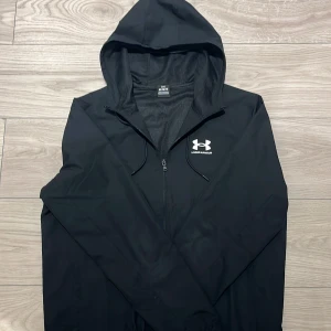 Svart vindjacka Under Armour - Svart vindjacka från Under Armour med huva och snörning. Jackan har dragkedja framtill och ett vitt logotryck på bröstet. Långärmad modell med elastiska muddar vid ärmslut. Perfekt för dig som gillar sportig och clean stil. 💨PRISET ÄR EJ HUGGET I STEN VID SNABB AFFÄRR 💨!!!