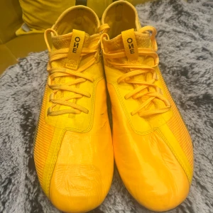 Gula Puma fotbollsskor med snörning elite - Säljer ett par riktigt snygga gula fotbollsskor från Puma med svart logga på hälen. Skorna har snörning, platt sula och rund tå. Ovandelen är tillverkad i syntetmaterial med mesh-detaljer på sidorna för extra ventilation. Perfekt för dig som vill sticka ut på planen.och är bara använd en gång 