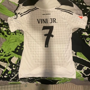 Real Madrid Vini Jr. 7 matchtröja M - Säljer en officiell Real Madrid fotbollströja från Adidas i storlek M, med VINI JR. och nummer 7 på ryggen. Tröjan är vit med subtilt hundtandsmönster, svarta detaljer och klubbmärke på bröstet. Materialet är lätt och andas, perfekt för match eller träning.