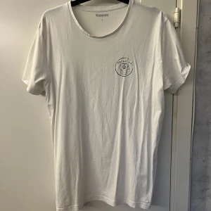 Vit t-shirt från Kappahl med tryck - Vit t-shirt från Kappahl i storlek L, med rund hals och korta ärmar. På bröstet finns ett litet tryck med texten 'PRINT UF' och en grafisk illustration. Den är i ett bra skick. Hör av er om ni har frågor!