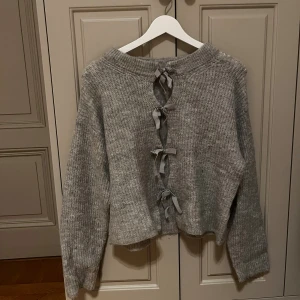 Grå stickad tröja med knyt från Gina Tricot - Supermysig grå stickad tröja från Gina Tricot med långa ärmar och snygga knytband längs ryggen. Tröjan har en loose passform och ribbad struktur som gör den extra skön. Perfekt för dig som gillar detaljer och vill ha något unikt i garderoben.