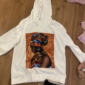 Vit hoodie med färgstark tryck - Säljer en vit hoodie med snörning i huvan och ett stort, färgstarkt tryck på bröstet som föreställer en kvinna med mönstrad sjal i blått, orange och grönt. Hoodien har lång ärm och avslappnad passform, perfekt för dig som gillar statement-plagg.