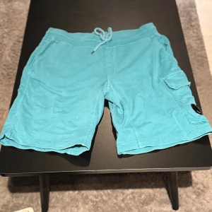 Turkosa shorts från C.P. Company - Säljer ett par turkosa shorts från C.P. Company med snörning i midjan och en praktisk cargoficka på sidan. Ikonisk linsdetalj på benet. Sköna och lätta, perfekta för varma dagar. Tillverkade i mjuk bomull.