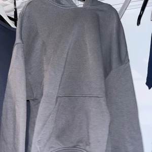 Grå hoodie med huva och ficka - En enkel grå hoodie med huva och stor magficka. Tröjan har en loose passform och är långärmad, perfekt för chill dagar. Materialet känns mjukt och är troligen bomull eller polyester. Snygg att matcha med jeans eller joggers.