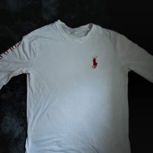 Vit långärmad tröja Polo Ralph Lauren - Vit långärmad tröja från Polo Ralph Lauren med rött broderat logotyp på bröstet och tryckt text längs ena ärmen