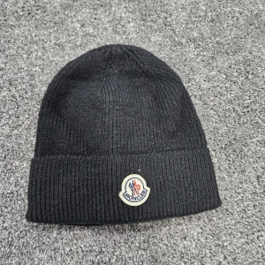 Svart ribbstickad mössa från Moncler - Svart ribbstickad mössa från Moncler med klassisk uppvikt kant och broderad logotyp framtill. Perfekt för kalla dagar och enkel att matcha med olika outfits. Tillverkad i mjukt material som håller dig varm och snygg.