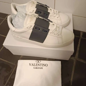 Valentino Garavani sneakers vit/grå - Snygga sneakers från Valentino Garavani i vitt skinn med bred grå panel över snörningen. Platt sula och rund tå, samt ikoniska nitar baktill på hälen. Kommer med originalkartong, dustbag och allting utom kvitto. Perfekt för dig som gillar exklusiv streetstyle.