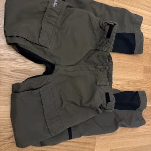 Lundhags gröna täckbyxor med svarta paneler - Säljer ett par gröna täckbyxor från Lundhags med svarta förstärkta paneler på knän och bak. Byxorna har stora fickor med lock, justerbar midja med kardborre och slitstarka material. Perfekta för utomhusaktiviteter och äventyr.