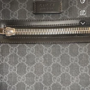 Gucci axelväska med GG-mönster - Snygg axelväska från Gucci i mörkgrått med klassiskt GG-mönster över hela väskan. Väskan har en silverfärgad dragkedja framtill och en bred axelrem i blått och rött. Materialet är polyester med detaljer i skinn. Perfekt för dig som vill ha en stilren och ikonisk väska.