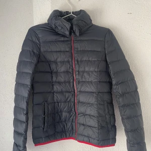 Svart pufferjacka från Esprit - Svart pufferjacka från Esprit med röda detaljer och dragkedja framtill. Jackan har hög krage, två fickor med knappstängning och quiltad design. Materialet är syntet och jackan har långärmade ärmar. Perfekt för kyliga dagar. Den har hål som är ihopsydda