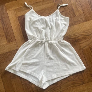 Vita shorts-jumpsuit från H&M Divided XS - Supermjuk vit jumpsuit med shorts från H&M Divided. Tunt frottéliknande material i 59% polyester och 41% bomull, med smala axelband som knyts och resår i midjan. Perfekt för varma dagar och chill vibes.
