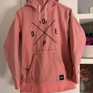 Dope jacka - Säljer en snygg rosa hoodie från Dope med svart grafiskt tryck på bröstet. Tröjan har huva med vita snören, magficka och ribbade muddar. Perfekt för dig som gillar streetwear och vill sticka ut lite extra.