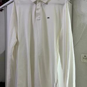 Vit långärmad skjorta Tommy Jeans XL - Vit långärmad skjorta från Tommy Jeans i storlek XL. Klassisk design med krage och knappar framtill, samt diskret logga på bröstet. Skjortan är gjord i mjuk bomull och har en clean look som funkar till många stilar.