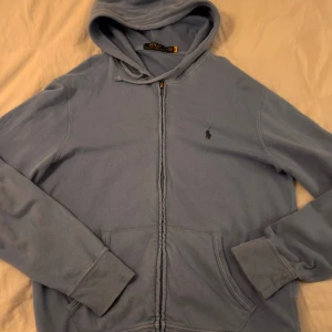 Ljusblå hoodie från Polo Ralph Lauren - Säljer en ljusblå hoodie från Polo Ralph Lauren med dragkedja och klassisk huva. Tröjan har långa ärmar, känguruficka och den ikoniska broderade loggan på bröstet. Perfekt för en chill och stilren look.