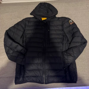Svart dunjacka Parajumpers med huva - Säljer en svart dunjacka från Parajumpers med huva och dragkedja framtill. Jackan har ett broderat märke på ena ärmen och quiltad design. Perfekt för kalla dagar och har en snygg, sportig look.