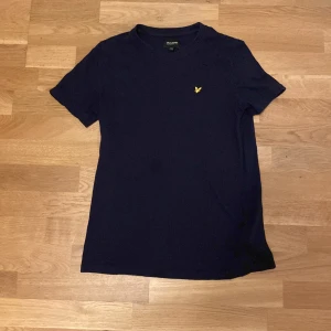 Mörkblå t-shirt från Lyle & Scott - Snygg mörkblå t-shirt från Lyle & Scott Junior med klassisk rund hals och korta ärmar. På bröstet sitter den ikoniska gula fågeln broderad. T-shirten är gjord i mjuk bomull och har en clean, stilren look som funkar till jeans eller shorts.