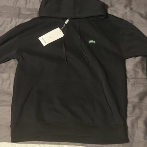 Svart hoodie från Lacoste med logga - Svart hoodie från Lacoste med klassisk grön krokodillogga på bröstet. Tröjan har huva med snörning, stor magficka och ribbade muddar. Perfekt för en chill och stilren look. Materialet är mjukt och känns skönt mot huden.                        📦 1-3 dagar ✅Tar emot bud.       Hoodien är lite liten i storlek så skulle kunna passa M