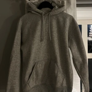 Grå hoodie med huva och ficka - Säljer en klassisk grå hoodie med huva och dragsko samt en stor magficka framtill. Tröjan har långa ärmar och ribbade muddar vid ärmslut och nederkant. 