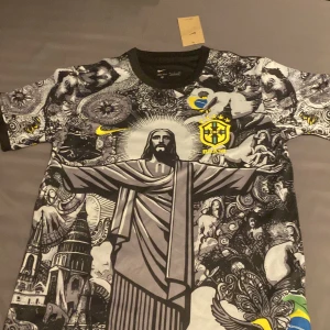 Nike Brasil Dri-Fit fotbollströja S - Unik Brasilien fotbollströja från Nike med svartvit konstnärlig grafik, Kristusstatyn och detaljer i gult och blått. Tillverkad i Dri-Fit material som andas. Korta ärmar och rund hals. Perfekt för dig som vill sticka ut på planen eller läktaren.