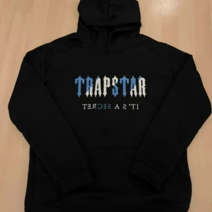 Trapstar svart hoodie med blå text - Svart hoodie från Trapstar med stor blå och vit logga på bröstet och texten 'IT'S A SECRET' under. Klassisk känguruficka framtill och huva. Perfekt för dig som gillar streetwear och vill sticka ut med en fet logga. Köpt utomlands alltså är det inte en äkta vara. Men är väldigt snygg.