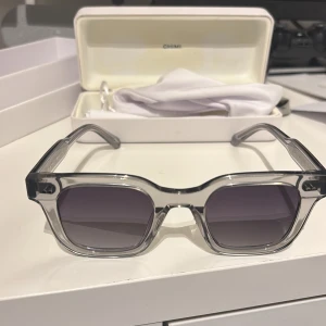 Grå transparenta solglasögon från CHIMI - Snygga solglasögon från CHIMI med fyrkantig form och transparent grå båge. Glasen är mörktonade och bågarna har en cool, modern look. Kommer med originaletui och putsduk. Perfekta för dig som vill sticka ut med stilrena accessoarer. Box till glasögonorna, box till boxen och en nk påse ingår. Kvitto följer också med. De är knappt använda. Pris kan diskuteras.