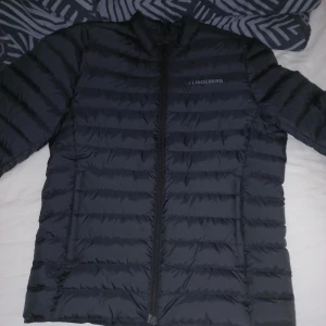 Svart dunjacka från J.Lindeberg - Snygg svart dunjacka från J.Lindeberg i storlek M. Jackan har ett stilrent, quiltat mönster med horisontella sömmar och hel dragkedja framtill. Perfekt för kyliga dagar och har en modern, normal passform. Diskret logga på bröstet.