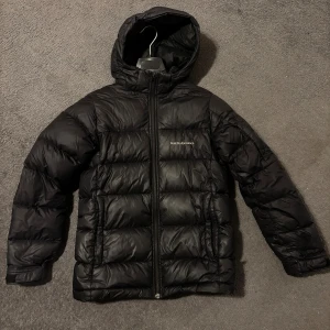 Svart Peak Performance dunjacka - Svart dunjacka från Peak Performance med huva och dragkedja framtill. Jackan har puffad design och logga på bröstet. Perfekt för kalla dagar och har en stilren look som funkar till det mesta. Jackan är långärmad och har praktiska fickor på sidorna. Det är storlek 160 och det finns en liten defekt på höger armbåge men annars är den i väldigt bra skick. 