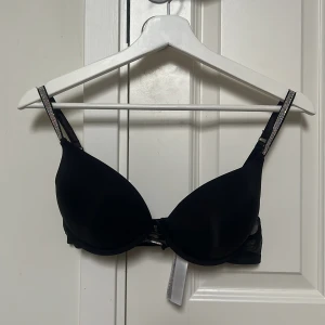 Svart push-up bh med strassdetaljer - Svart push-up bh med glittrande strass på axelbanden och mellan kuporna. Axelbanden är justerbara och bakstycket har meshdetaljer. Materialet är mjukt och slätt. Perfekt för dig som gillar glam och vill ha något extra.