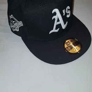 Svart New Era 59FIFTY Oakland A's keps - Säljer en svart New Era 59FIFTY keps med Oakland A's logga broderad i vitt framtill. Kepsen har platt skärm, broderade detaljer på sidorna och MLB-logga bak. Insidan har klassisk 59FIFTY-märkning och grå underskärm. Perfekt för dig som gillar sportig streetstyle.