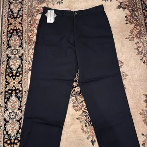 Navy blå chinos från Dockers, 36x34 - Navy blå chinos från Dockers i klassisk rak modell med bälteshällor och knappstängning. Snyggt stilrena och tillverkade i mjukt bomullsmaterial. Passar dig som gillar enkel och clean look. Taggen sitter kvar på byxan.