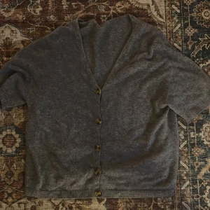 Grå kofta med kort ärm och knappar - Mysig grå kofta med korta ärmar och snygga bruna knappar framtill. Koftan har v-ringning och är stickad i ett mjukt material, perfekt att slänga på sig över en t-shirt eller linne. Enkel och stilren design som funkar till det mesta.