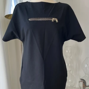 Svart kortklänning med dragkedja - Svart kortklänning med rak passform och korta ärmar. Framtill finns en cool silverfärgad dragkedja som detalj över bröstet. Klänningen har fickor i sidorna och är tillverkad i ett mjukt bomullsmaterial. Perfekt för dig som gillar minimalistisk och stilren design.