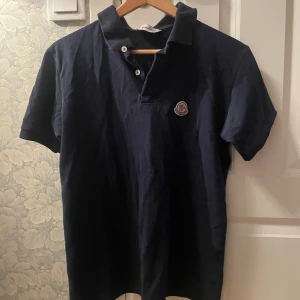 Mörkblå Moncler pikétröja - Snygg mörkblå pikétröja från Moncler med klassisk krage och två vita knappar framtill. Tröjan har Moncler-logga på bröstet och är tillverkad i mjuk bomull. Perfekt för dig som gillar stilrena och exklusiva plagg.