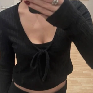 Svart croppad kofta med knyt - Säljer en jätte söt lite kortare kofta med ribbad struktur och långa ärmar. Toppen har en snygg v-ringning och knyt framtill som ger en trendig look. Perfekt att styla med höga jeans eller kjol. Aldrig använd❤️