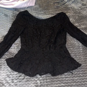 Svart spetsblus med volang - Säljer en svart blus med lång ärm och vacker spets över hela toppen. Blusen har en feminin volang nedtill som ger en snygg siluett. Perfekt för dig som gillar romantiska detaljer och vill ha något extra till din outfit.