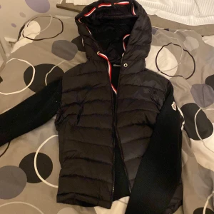 Byta en cardigan med det här - Svart pufferjacka från Moncler med stickade ärmar och huva. Jackan har dragkedja framtill och coola röd-vit-blå detaljer på snörningen. Moncler-logga på ärmen och quiltad kropp ger en snygg och sportig vibe. Perfekt för dig som gillar streetstyle.