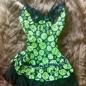 Grön blommig topp med spets - Supersöt topp med grönt och vitt blommönster på svart botten, spetsdetaljer upptill och volangkant nertill. Axelbandslös look med smala axelband och liten rosett framtill. Perfekt för dig som gillar färg och retrovibbar!