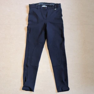 Mörkblå ridbyxor från CRW, stl 38 - HELT OANVÄNDA RIDBYXOR Snygga mörkblå ridbyxor från CRW med smal passform och förstärkta knäpartier. Byxorna har bälteshällor, knapp och dragkedja framtill samt CRW-logga vid linningen. Perfekta för dig som gillar ridning och vill ha en sportig look. Kardborre längst ner på byxbenen 