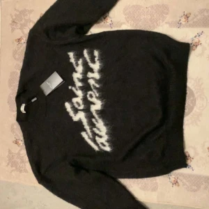 Svart stickad Saint Laurent-tröja - Svart stickad tröja från Saint Laurent med vit, suddig logotyp framtill. Tröjan har rund halsringning och långa ärmar. Mjuk och fluffig känsla, perfekt för att lyfta din outfit med en lyxig touch.