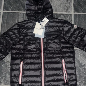 Svart Moncler dunjacka med huva - Svart dunjacka från Moncler med glansig finish, huva och två bröstfickor med knapp. Jackan har röda och vita detaljer på dragkedjorna och klassisk Moncler-logga på ärmen. Perfekt för kalla dagar och har en snygg quiltad look.