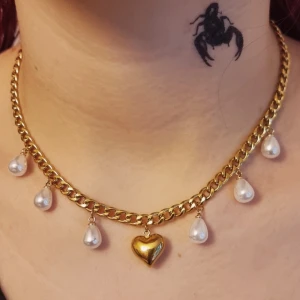 Guldfärgat halsband med pärlor och hjärta - Chunky guldfärgat halsband med droppformade vita pärlor och ett glänsande hjärthänge i mitten. Material-rostfritt stål,  kedjetjocklek-5cm.  Nikelfria.