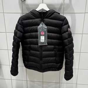 Svart Colmar pufferjacka med huva - Svart pufferjacka från Colmar Originals med huva och två sidofickor med dragkedja. Jackan har ett stilrent, quiltat mönster och är tillverkad i syntetmaterial. Perfekt för dig som vill ha en snygg och funktionell jacka med cool streetkänsla.