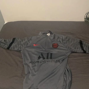 Grå PSG långärmad fotbollströja Nike - Säljer en grå långärmad PSG fotbollströja från Nike med svarta detaljer och röd Nike-logga. Tröjan har mönstrade ärmar och PSG-emblem på bröstet. Materialet är lätt och andas, perfekt för träning eller match. Snygg design med Accor Live Limitless-tryck framtill.