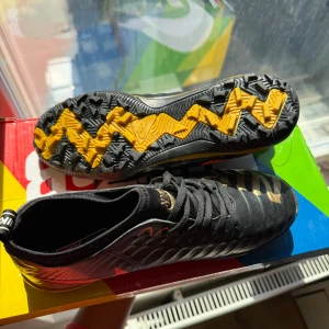 Nike fotbollsskor svart/gul/orange - Säljer ett par Nike fotbollsskor med svart ovandel, orange swoosh och gul detalj på hälen. Sulan har ett coolt mönster i svart och gult med grovt grepp. Skorna har snörning och texten NIKE i guld på tån. Materialet är syntet och skinn för bästa känsla på planen.