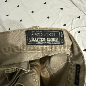 Beige cargopants från Angelo Litrico - Säljer ett par beige cargopants från Angelo Litrico med raka ben och flera fickor på sidorna. Byxorna har klassisk knapp och dragkedja framtill samt markerade sömmar över knäna för en cool look. Perfekt för dig som gillar streetstyle.