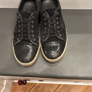 Svarta sneakers från Lanvin med mocka - Svarta sneakers från Lanvin med ovandel i mocka och glansig tå i skinn. Klassisk rund tå och platt sula i beige. Skorna har snörning med svarta skosnören och metallöglor. Snygg kontrast mellan material och färg, perfekt för dig som gillar stilrena detaljer. Allt og medföljer. OBS! pris ej hugget i sten 