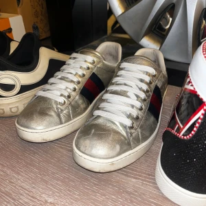 Gucci sneakers i silver och svart skinn - Säljer ett par feta äkta Gucci sneakers i metallic silver skinn med klassiska röd-blå ränder på sidorna. Baksidan har svart ormskinnsdetalj med Gucci-logga. Vita snören och vit sula. Perfekt för dig som vill sticka ut med lyxiga detaljer och cool design.