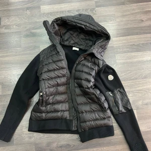Moncler cardigan - Svart pufferjacka från Moncler med stickade ärmar och quiltad kropp. Jackan har en stor huva, dragkedja framtill och fickor med dragkedja på ärmen. Moncler-logga på vänster ärm. Snygg och modern look med blandat material.