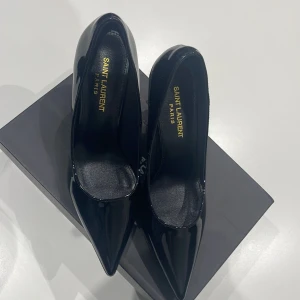 Svarta Saint Laurent pumps med YSL-klack - Säljer ett par svarta pumps från Saint Laurent i lackat skinn med spetsig tå och hög klack i guld med YSL-logga. Skorna har en elegant och modern siluett som verkligen sticker ut. Perfekta för dig som vill ha något extra till outfiten.